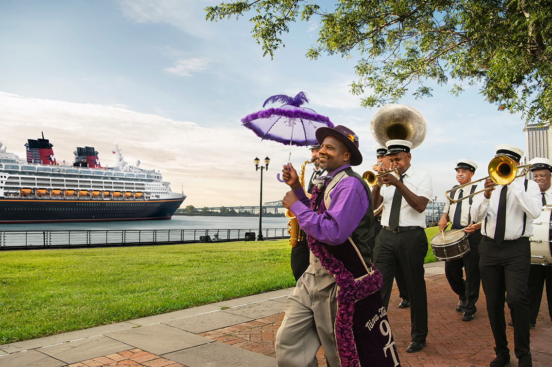 Un homme vêtu d'une tenue joyeuse et tenant un parasol duveteux conduit un petit groupe de musiciens devant un bateau de croisière Disney
