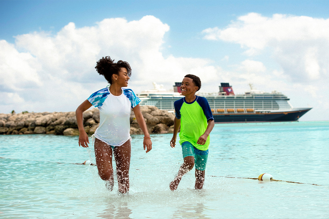 Un garçon et une fille courent le long du rivage sur Disney Castaway Cay avec un navire de la Disney Cruise Line en arrière-plan